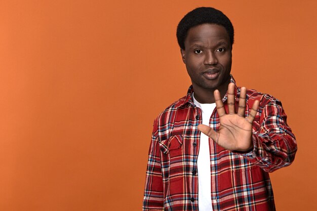 Confident serious young dark skinned man showing stop sign, lifting open palm and saying No. Stylish African guy making refusal warning gesture with hand in front of him, expressing refusal