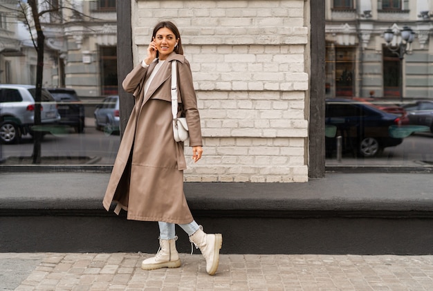 Confident short haired woman wearing casual beige color coat and white textured leather shoulder bag, walking in street of European city