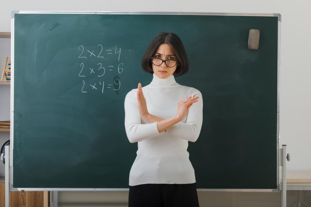 자신감 있는 제스처를 보여주는 젊은 여교사는 안경을 쓰고 칠판 앞에 서서 교실에서 글을 씁니다.