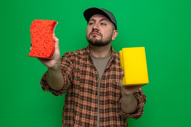 Confident slavic cleaner man holding and looking at sponges 