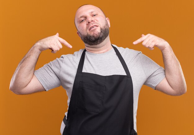 Confident slavic middle-aged male barber in uniform points at himself isolated on orange wall