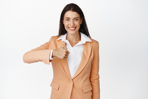 親指を立てて示す自信を持って笑顔のビジネス女性は、白い背景に対してスーツに立っている何か良いよくやったジェスチャーを承認します
