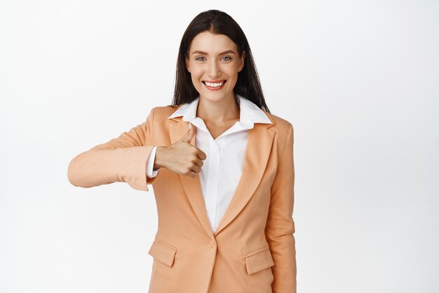 Уверенно улыбающаяся деловая женщина, показывающая большой палец вверх, одобряет что-то хорошее, хорошо сделанный жест, стоя в костюме на белом фоне