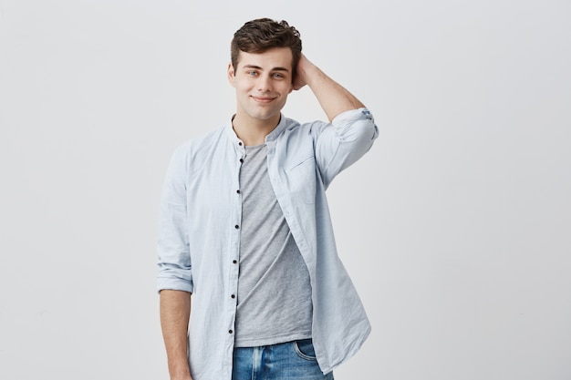 Confident smiling caucasian man, with dark hair and appealing blue eyes, looking  with pleased expression, dressed in blue shirt over t-shirt, holding his hand behind the back of his head.