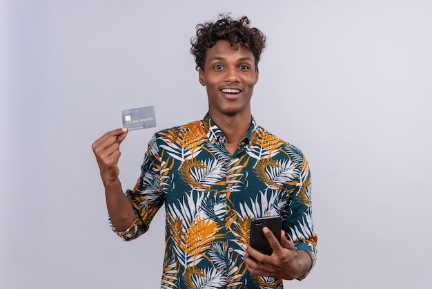 Уверенный и улыбающийся симпатичный темнокожий мужчина с вьющимися волосами в рубашке с принтом листьев показывает кредитную карту, держа в руке мобильный телефон