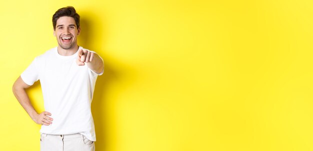 Confident smiling man pointing at you camera standing in white clothes against yellow background