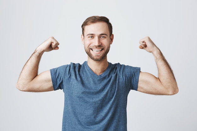 Уверенный улыбающийся человек тренируется в тренажерном зале, сгибает сильные бицепсы, показывает мышцы