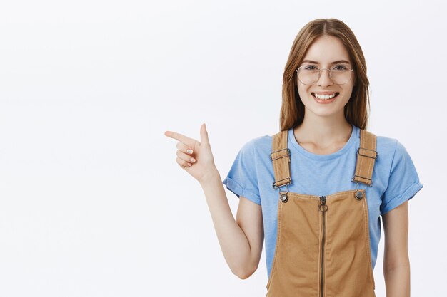 Confident smiling pretty girl student in glasses looking happy, pointing finger left at your logo