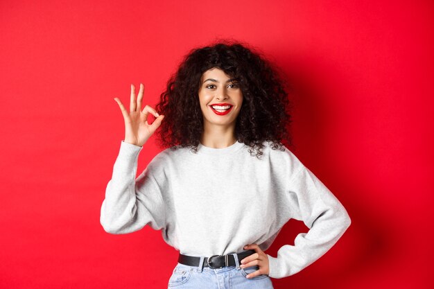 Confident smiling woman in casual clothes, showing OK sign to approve or like product, guarantee good quality, making a compliment, red background.