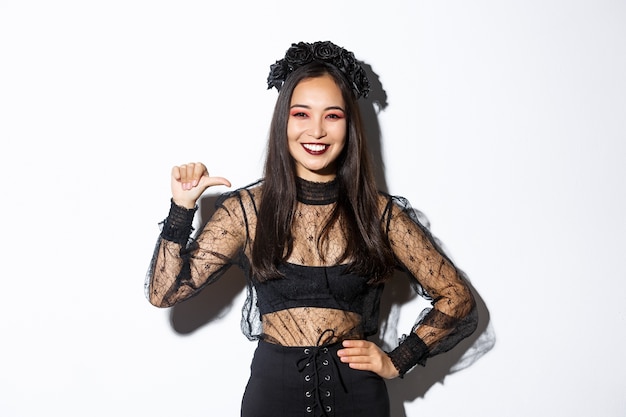 Confident smiling woman feeling like professional, pointing at herself while standing over white background in black gothic dress for halloween party.