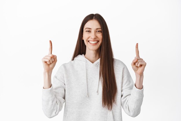 Confident smiling woman recommending logo click on website banner pointing fingers up and looking at camera showing best deal in store standing over white background