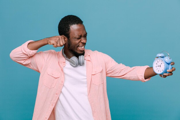 Confident standing on fighting pose holding alarm clock young africanamerican guy wearing headphones on neck isolated on blue background