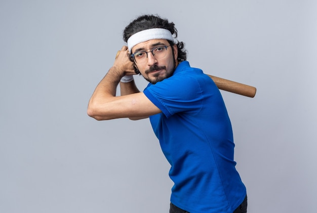 Confident standing in fighting pose young sporty man wearing headband with wristband holding baseball bat 