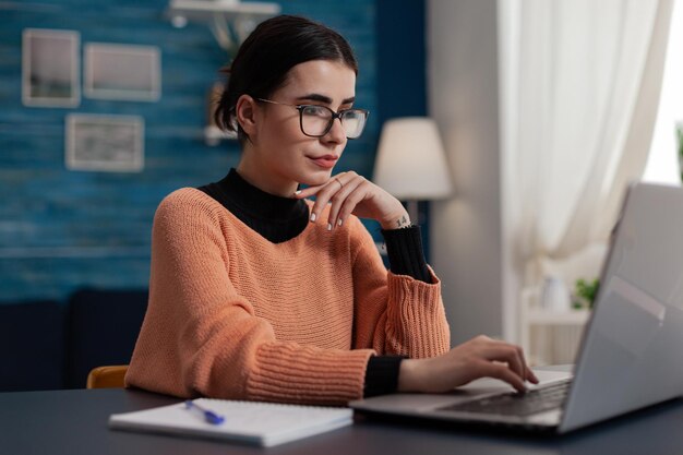 Confident student studying remote looking at laptop screen. Content creator with glasses casually typing sitting at home desk. College girl browsing the internet. Freelancer working from home sending