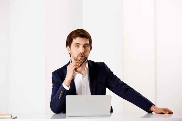 Confident, stylish businessman in suit, sit office desk near laptop, looking thoughtful