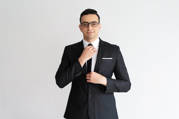 Confident stylish handsome young businessman adjusting necktie and looking at camera.