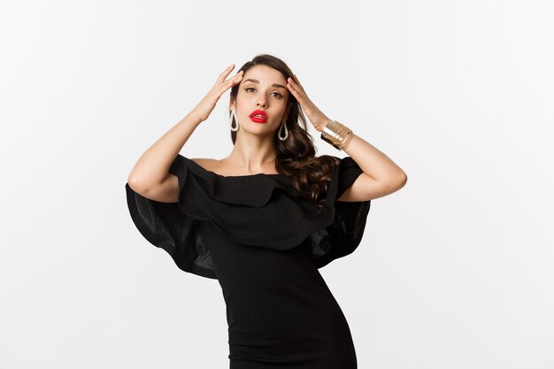 Confident and stylish woman in black elegant dress, looking sassy at camera, standing over white background.