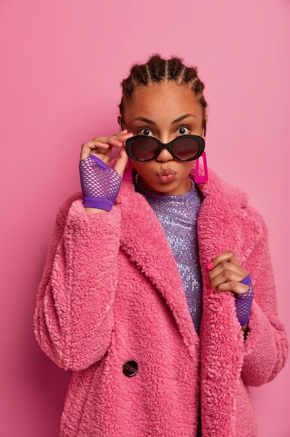 Confident stylish woman keeps lips rounded, flirts with lover, wears sunglasses and warm coat of last fashion trend, looks with widely opened eyes, poses against pink wall. Glamour and style