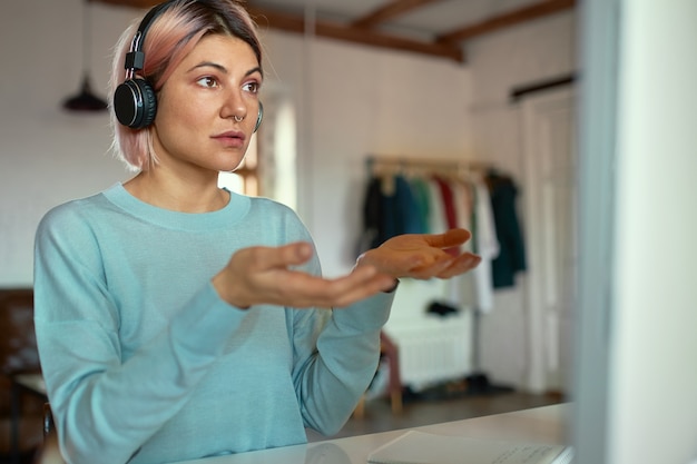 Confident stylish young pink haired woman wearing wireless headphones and nose ring gesturing emotionally while conducting webinar via video conference chat on cumputer.