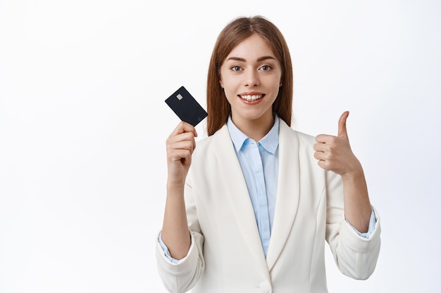 Confident, successful business woman shows plastic credit card and thumbs up, smiling pleased, recommend bank, standing over white wall