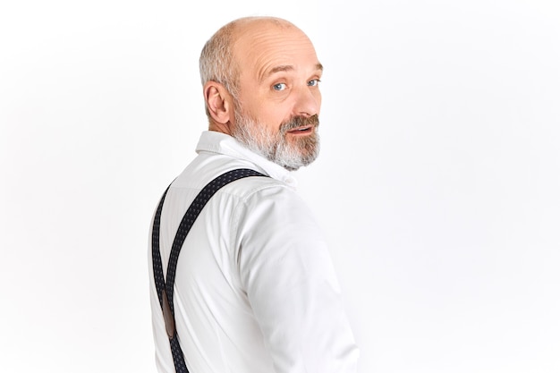 Confident successful European man on retirement in stylish white shirt and suspenders turning round and looking at camera with serious facial expression. People, age, maturity and elegance concept