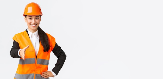 Уверенная в себе успешная женщина-архитектор-лидер строительства в защитном шлеме со светоотражающей курткой протягивает руку для рукопожатия, приветствуя деловых партнеров на строительной площадке, стоящей на белом фоне