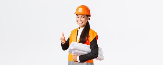 Confident successful female asian construction manager architect in safety helmet and jacket showing thumbup and carry blueprints of building project guarantee quality white background