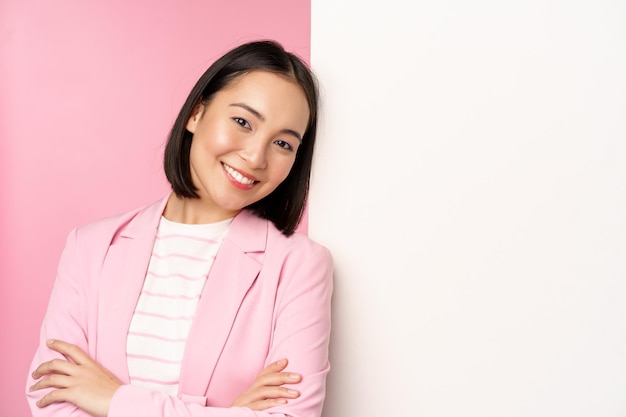 Confident successful japanese office lady in suit cross arms looking as professional at camera leaning on white wall with advertisement empty copy space for logo pink background