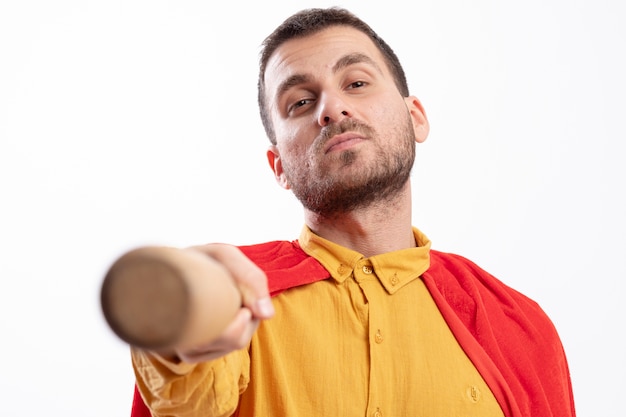 Confident superhero man with red cloak holds out baseball bat isolated on white wall