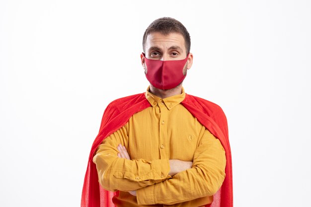 Уверенный супергерой в красном плаще в красной маске стоит со скрещенными руками, изолированными на белой стене