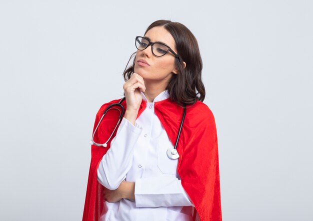 Confident superwoman in doctor uniform with red cape and stethoscope in optical glasses puts hand on chin and looks at side isolated on white wall