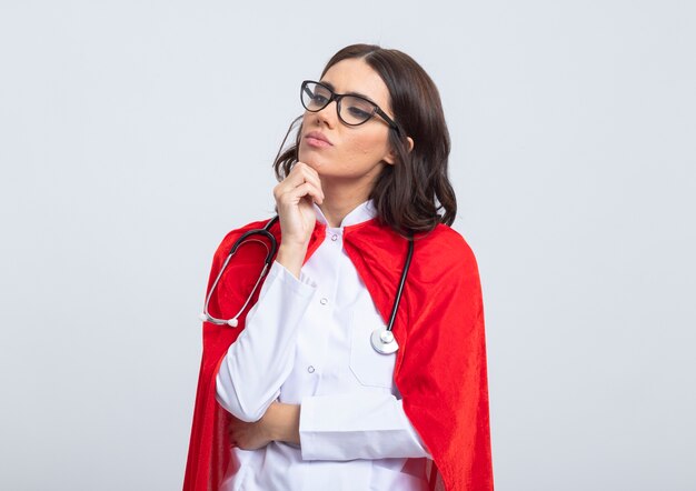 赤いマントと光学メガネの聴診器で医者の制服を着た自信のあるスーパーウーマンは、あごに手を置き、白い壁で隔離された側を見ます