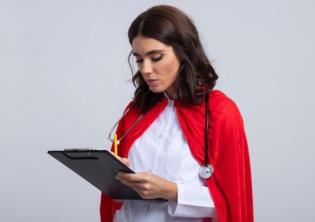 Confident superwoman in doctor uniform with red cape and stethoscope writes on clipboard with pencil isolated on white wall
