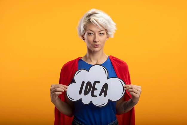 Confident superwoman with red cape holds idea bubble isolated on orange wall