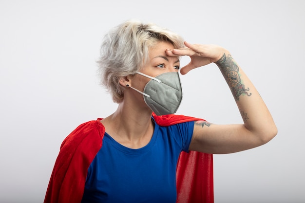 Confident superwoman with red cape wearing medical mask keeps palm at forehead looking at side isolated on white wall