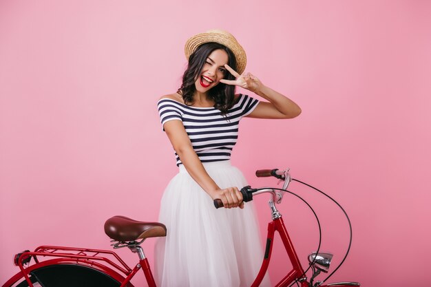 Уверенно загорелая женщина позирует с велосипедом и выражает счастье. Крытая фотография веселой девушки в романтическом наряде.