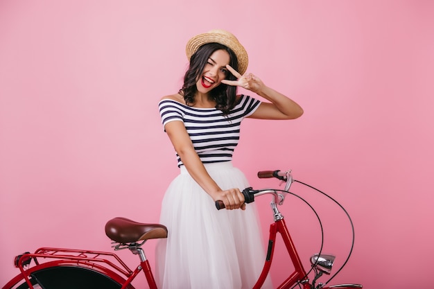 Confident tanned woman posing with bicycle and expressing happiness. Indoor photo of debonair girl in romantic outfit  having fun.