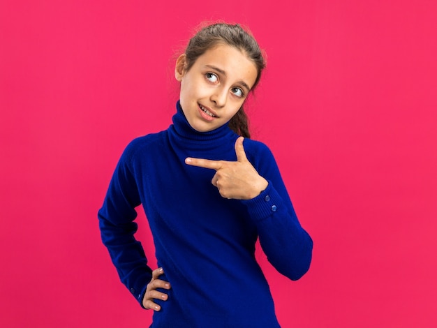 Confident teenage girl keeping hand on waist looking and pointing at side isolated on pink wall with copy space