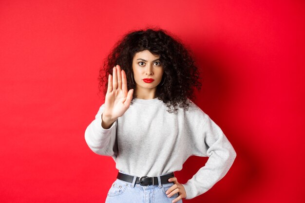 Confident tensed woman extend hand to say stop, disapprove action and prohibit it, make no gesture, standing on red background and forbid something.