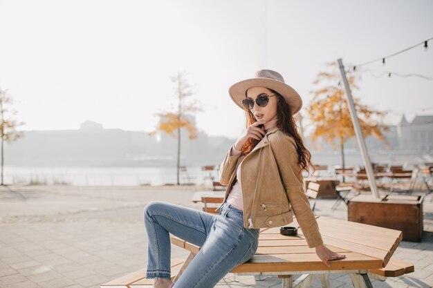 Confident woman in denim pants sitting on table in street cafe