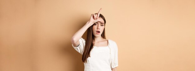 Confident woman mocking people with loser sign being mean standing on beige background