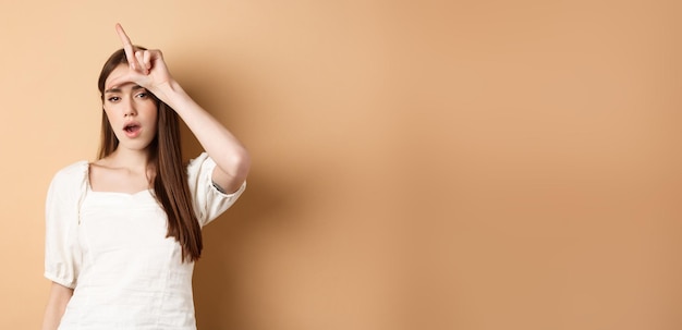 Free photo confident woman mocking people with loser sign being mean standing on beige background