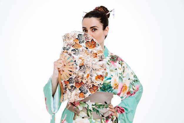 Confident Woman in traditional japanese kimono holding hand fan coning mouth with it on white