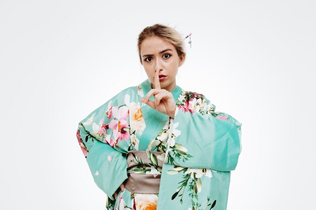 Уверенная женщина в традиционном японском кимоно делает жест молчания с пальцем на губах на белом