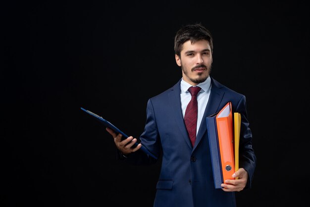 Confident young adult in suit holding several documents on isolated dark wall