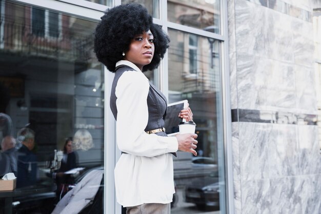 Confident young african businesswoman standing outside the office holding digital tablet and disposable coffee cup