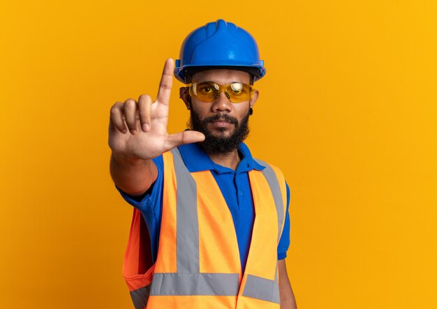 Уверенный молодой афро-американский строитель человек в защитных очках в униформе с защитным шлемом, направленный вверх, изолированный на оранжевой стене с копией пространства