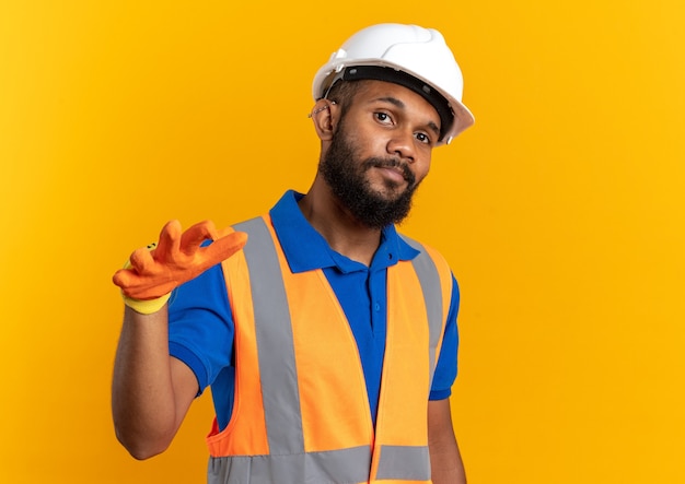 Уверенный молодой афро-американский строитель человек в форме с защитным шлемом и перчатками держит руку открытой изолированной на оранжевом фоне с копией пространства