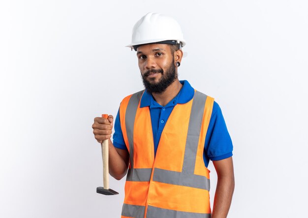 Confident young afro-american builder man in uniform with safety helmet holding hammer upside down isolated on white wall with copy space