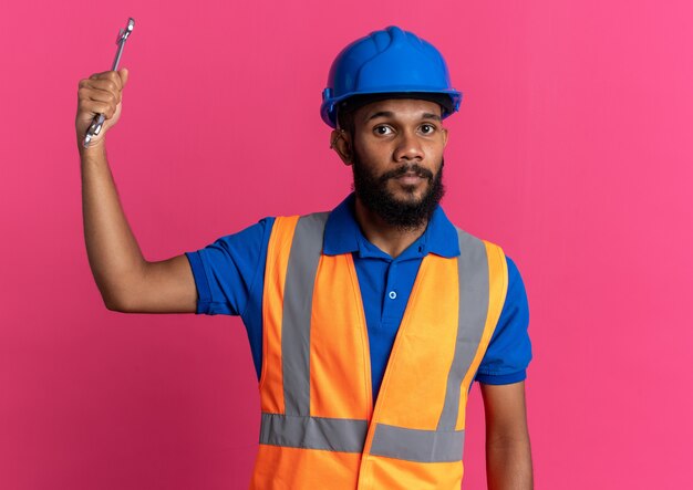 Уверенный молодой афро-американский строитель человек в форме с защитным шлемом, держащий ключ мастерской, изолированный на розовом фоне с копией пространства